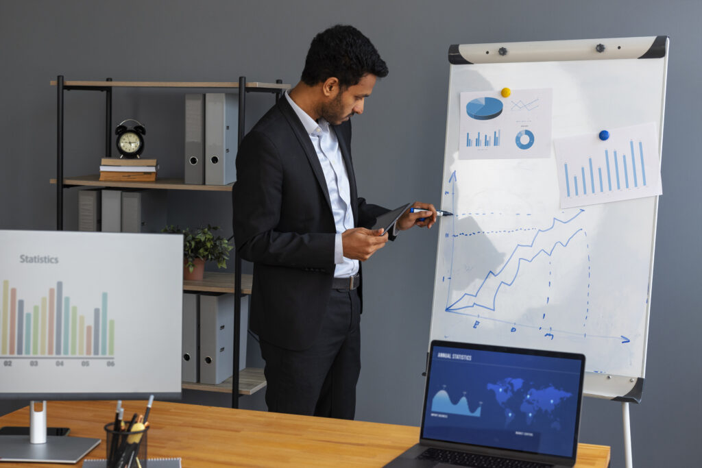 A man in a formal suit stands beside a whiteboard with charts and graphs, pointing at data with a pen while holding a tablet. The office setting includes a desk with a laptop and monitor displaying statistics, and a shelf with files and a clock in the background.
